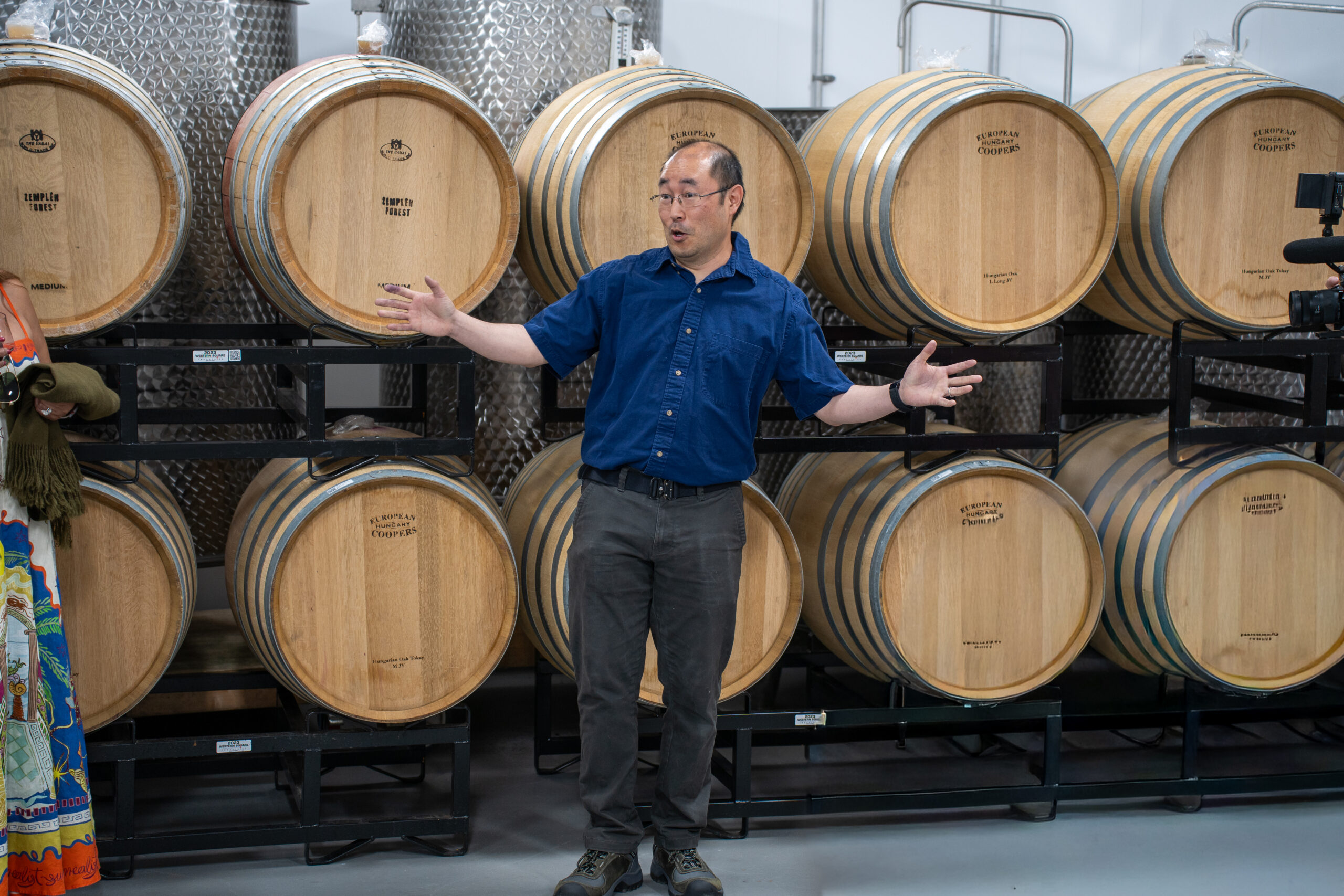 Winemaker in blue button down in front of wine barrels talking animated to people who are listening
