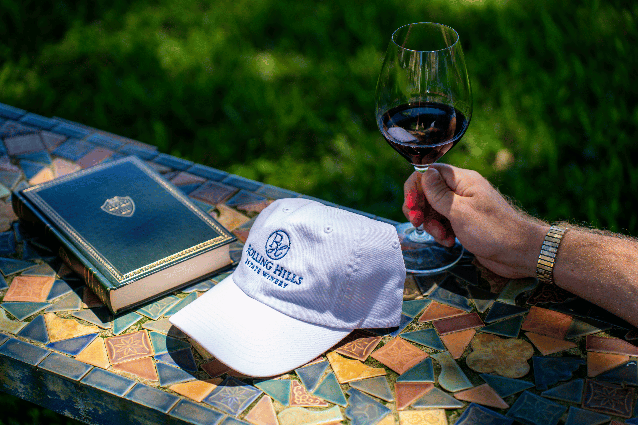 Outside table with a white rolling hills baseball hat, a green book and a man's hand holding a glass of red wine