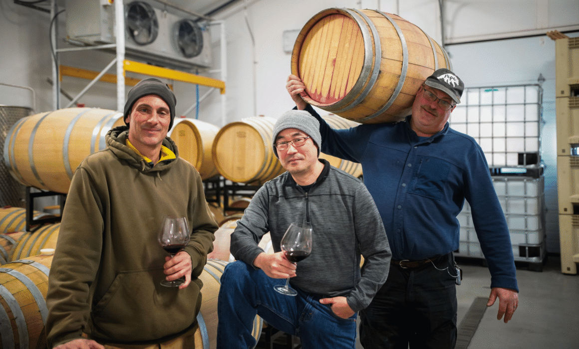 three men in winery. Two holding wine glasses with red wine and the other holding a wine barrel, all smiling
