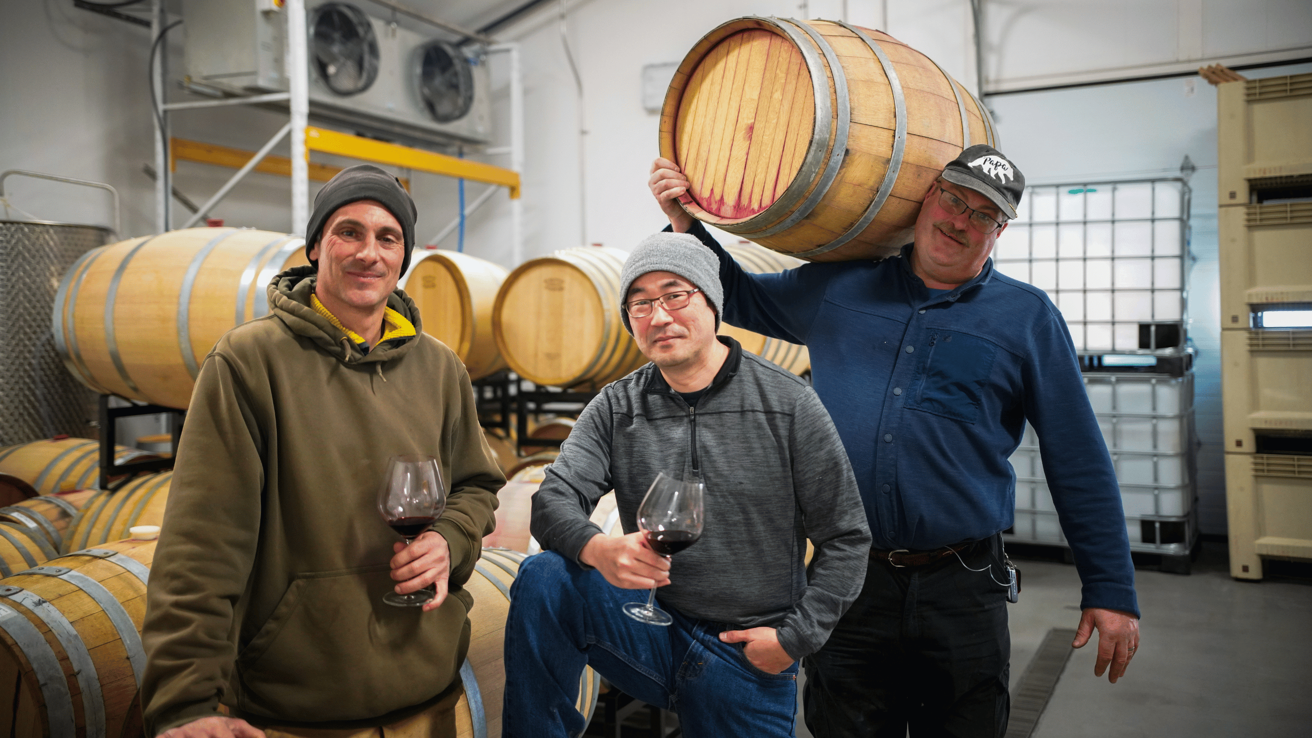 three men in winery. Two holding wine glasses with red wine and the other holding a wine barrel, all smiling